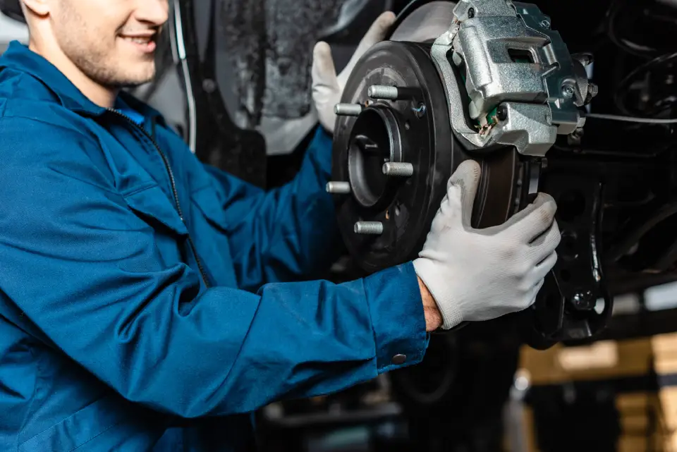 Mechanic in blue overalls replacing a car brake rotor and caliper, wearing gloves under the vehicle.