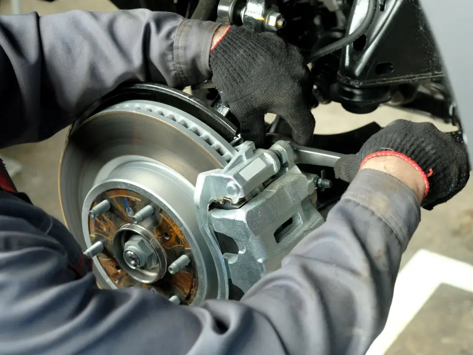 Close-up of a mechanic's gloved hands working on a car's brake rotor and silver caliper, with the wheel hub visible.